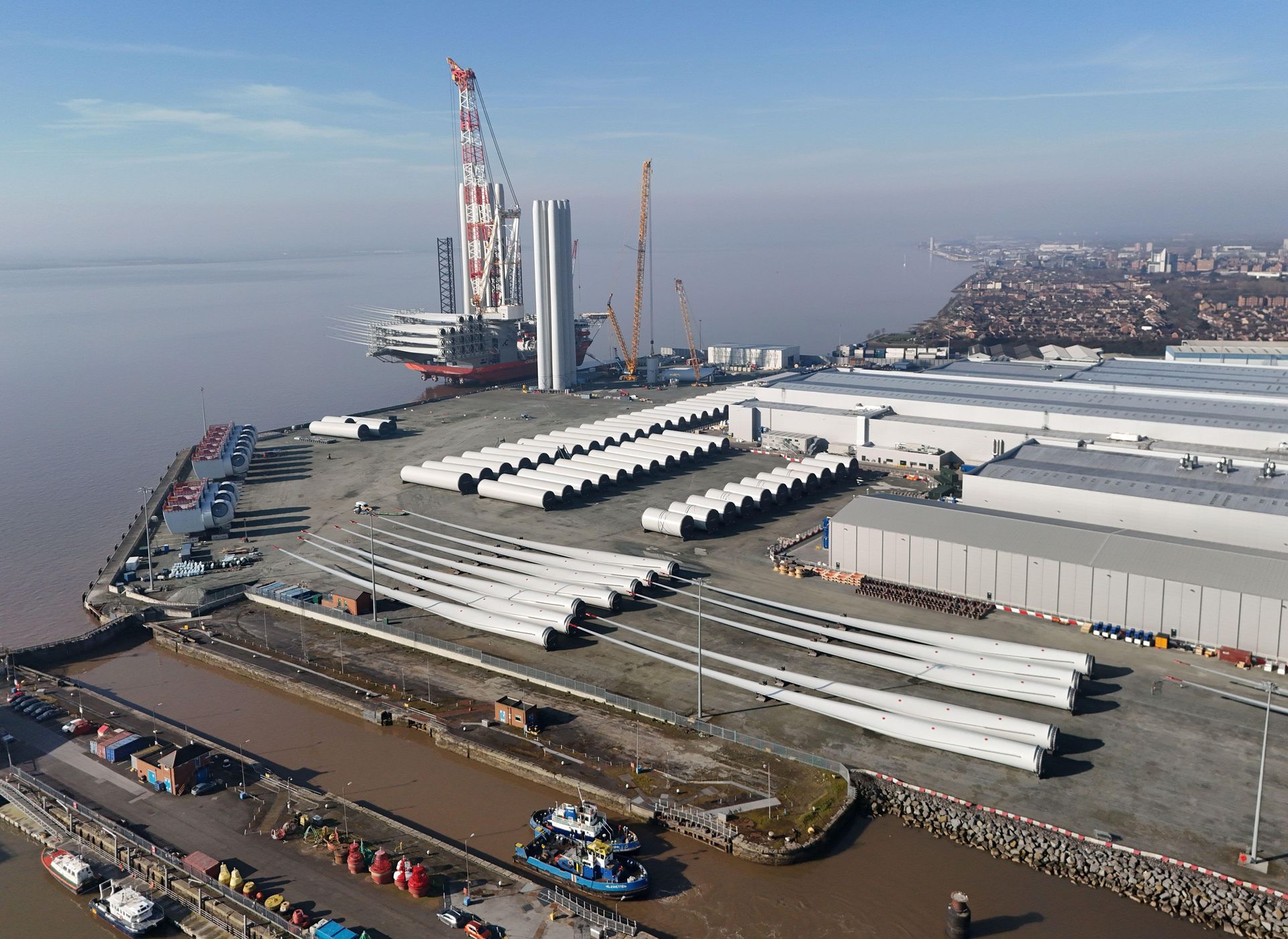 Race Cottam Associates Ltd — Aerial view of an industrial port with wind turbine blades stored in rows, large warehouses, and an offshore platform docked along the river beside a
