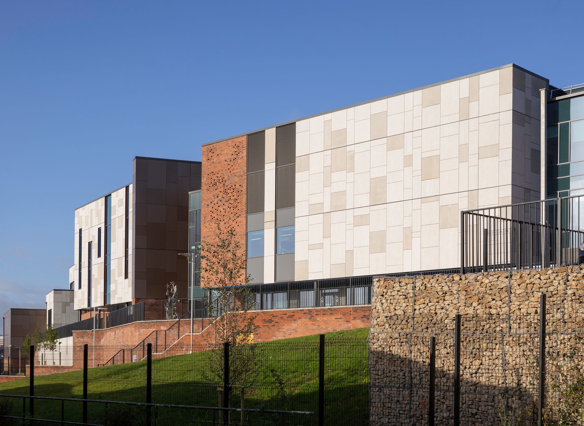 Race Cottam Associates Ltd — Exterior view of a new school building with a mix of light-coloured cladding panels, projecting brickwork detailing, set within a landscaped area with